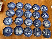 Photo showing all 19 Royal Copenhagen collector plates laid out on a wooden floor, depicting various winter and maritime scenes in blue and white.