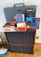 Overall set showing Sony speakers stacked, a box with tapes, Hitachi and Sony cassette recorders with carrying case, and VHS tapes.