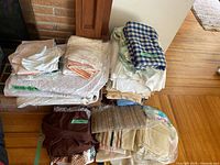 Stacked bundles of assorted linens on floor, showing variety of tablecloths, aprons, napkins, runners, placemats including blue checked fabric and brown aprons