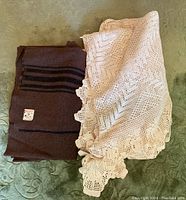 Brown wool blanket folded with lace bedspread on top on a green surface.