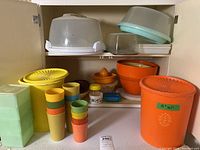 Photo showing a collection of vintage Tupperware items on white cabinet shelves including orange and yellow storage containers, a set of multicolored glasses, mixing bowls, citrus juicer, and other small containers.