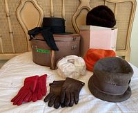 Overview of vintage hats, gloves, hat boxes on bed showing six hats including black wide-brimmed with scarf, orange, gray fur, white ruched, dark brown fur; red and brown gloves in front, and two hat boxes behind.