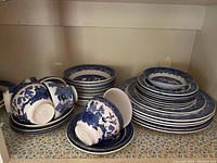 A shelf showing stacked Willow Pattern teacups and plates arranged in piles, some cups resting on saucers.