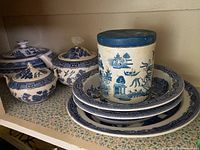 Photo showing multiple pieces of blue and white Willow pattern china including two covered pots, stacked plates, and a cylindrical canister with a blue lid.