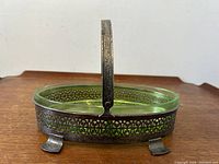 Side view of oval Vaseline glass condiment bowl with ornate silvered pierced metal holder and decorative raised handle.