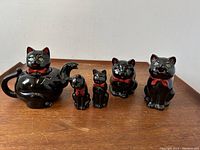Full view of the five-piece black cat redware tea set including teapot, creamer, salt shaker, pepper shaker with chipped ear, and sugar bowl with age crack.