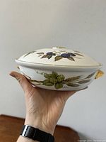 Side view of the circular lidded casserole showing floral and fruit design, held in hand for scale.
