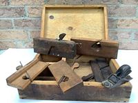 Photo showing the wooden storage box open with six antique wood planes inside, five wooden-bodied and one metal-bodied plane visible, surfaces showing heavy wear and patina.