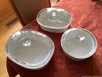 Photo showing all three white CorningWare casserole dishes with clear Pyrex glass lids on a table with red tablecloth.