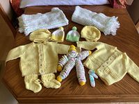 Full lot showing two yellow hand-knit baby sweaters, two yellow bonnets, one pair of yellow booties, two baby caps, a vintage knitted clown toy, and two fringed baby throws on a table