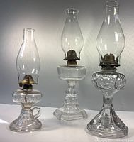 Three antique coal oil lamps in varying shapes and sizes displayed together on a white surface against a gray background. Each lamp has clear glass chimneys and brass burner mechanisms visible. The left lamp has a handle on the base, the center lamp has a hexagonal pedestal base, and the right lamp has an ornate pedestal with raised floral designs.