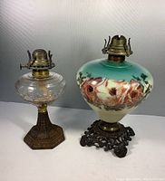 Two oil lamp bases displayed side by side showing front views. Painted milk glass lamp with floral design on right has ornate metal base. Clear glass lamp with brass burner on left has hexagonal metal base.