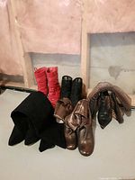 Five pairs of women's high heel boots arranged on floor, showing variety of colors and materials including black suede, shiny bronze, metallic bronze, red patent leather, and black quilted leather.
