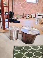 Two canning pots of different sizes with lids and handles, positioned on floor in room with unfinished walls and carpet.