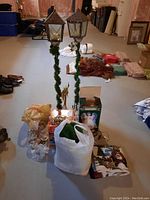 Photo showing the tall metal lantern candle holders wrapped in garland, Christmas figurines, ornaments, and bags containing other decorations on a basement floor.