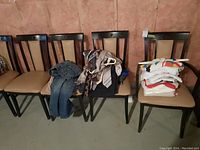 Photo showing four chairs with clothing items: denim pants on the first chair, assorted ties and scarves piled on the second chair, balloons or possibly more clothing on third chair obscured, folded shirts and t-shirts stacked on the fourth chair.