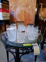 10 Bohemia crystal mugs arranged on a glass-topped table in a basement setting.