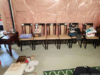 Six wood framed dining chairs with light brown vinyl seats and backs, arranged in a row in a basement with pink insulation wall.
