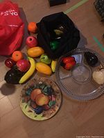 Photo showing assorted faux fruits and vegetables, a black bag containing some items, clear glass sectional plate with fruit shaped pieces, and a ceramic plate with fruit artwork