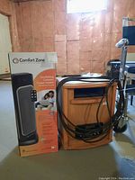 Photo showing Comfort Zone ceramic tower heater box next to Heat A Lot infrared heater with wooden casing and power cord wrapped around base in basement setting.