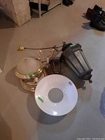 Top-down photo showing three round light fixtures: one bronze metal and glass lantern with textured glass panels, a polished metal and dome glass fixture, and a white glass reflector shade with a center hole.
