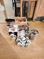 Overview photo with mugs, espresso cups with saucers stacked in front, stainless steel ice buckets behind, and boxed kitchen appliances.