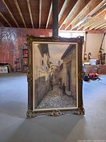 Full front view of framed painting showing street scene with buildings and person walking on cobblestone street.