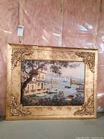 Framed coastal scene showing boats, water, a building, and a leafy tree with ornate gold-toned frame