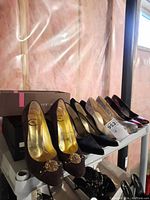 Five pairs of women's designer high heel closed toe shoes arranged on a white shelf with shoe boxes in the background. Shows variety of shoe styles and colors.