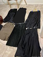 Seven skirts laid out on tile floor, mostly black with one olive skirt, showing varying styles and details.