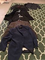 Seven men's suit jackets arranged in a row on a green patterned carpet, showing the variety and color of suits included in the lot