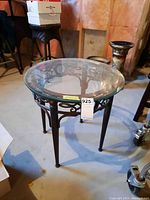Front and top view of round side table with clear glass top and decorative metal base on concrete floor in basement.
