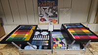 Full view of the opened Artist Loft art set showing the layout of colored pencils, pastels, paint tubes, brushes, palettes, and an instructional book.