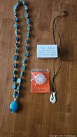 Photo showing two necklaces on a wooden surface: turquoise scarab bead necklace with large scarab pendant, white pendant necklace with black cord, handwritten note on 'Heir Agtau' traditional jewelry and a Bethlehem Jewelry tag.