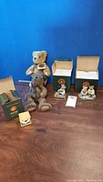 Full view of the 3 ceramic bear figurines with original boxes plus 5 plush teddy bears of varying sizes and colors on a wooden surface against a blue background