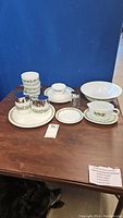 Photo showing a group of white ceramic dishware with green floral patterns including plates, bowl, gravy boat, creamer, sugar bowl, butter dish, and condiment servers arranged on wooden table.