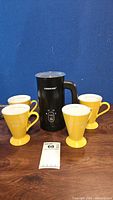 All five items together: 1 black Farberware electric frother with handle and 4 yellow Mary Kay Journey mugs.