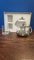 Full set with glass teapot, stainless steel top and handle, with infuser inside, and two clear glass mugs, all placed beside white foam packaging insert.