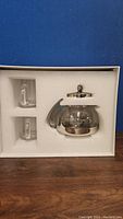 Photo showing glass teapot with stainless steel handle, lid, and infuser, plus two glass mugs in foam packaging box.