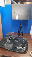 Side view of the black Gleam sheet music stand extended on tripod legs next to the black carrying case with logo.