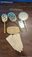Photo showing the front of two gold plated vintage hand mirrors, hairbrush with floral design, small gold mesh coin purse, gold comb, and beige gloves.