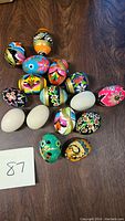Top view of 16 decorative eggs, 13 painted with colorful floral and geometric designs, and 3 unpainted plain eggs, arranged on a wooden surface.