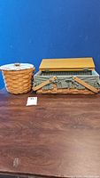 Two woven baskets placed side by side on a wooden surface against a blue background. Left basket is round with a plaid padded fabric lid and wooden knob; right basket is rectangular with a wooden hinged lid and two clasp straps.