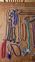 Photo showing multiple beaded necklaces in various colors including white, blue, purple, red, black, turquoise, and silver tones, also includes a purple tassel necklace and some silver-tone pendants and charms.