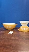 Three Pyrex Butterfly Gold mixing bowls stacked and side by side on a wooden surface against a blue background. The bowls have a golden yellow exterior with a white floral and butterfly pattern and white interior.