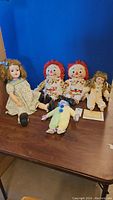 Shows lot of five dolls including Raggedy Ann & Andy, vintage girl doll in blue floral dress with bow, small cream pajama doll, and colorful small clown doll. Dolls are sitting on wooden surface against blue background.