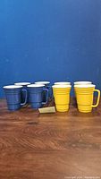 Set of eight ceramic mugs styled like Solo cups in two colors, six blue and two yellow, arranged on a wooden surface against a blue background.