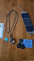 Photo showing necklace with Buddha pendant on dark beaded chain, wide blue beaded bracelet with silver accents, and round black beaded earrings attached to Blue River Designs card.