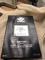 Front of black textured Creed Aventus box showing mounted silver label with horseman and “Creed” logo at bottom left.