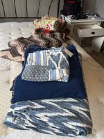 Photo showing storage of bedding items on mattress: includes two round floral throw pillows, a plaid wool blanket, a brown crocheted Afghan, a solid blue blanket, and a blue and white reversible comforter folded